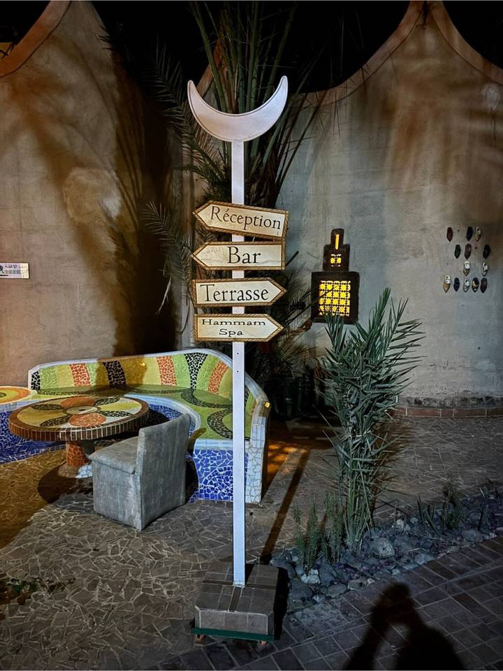 Rustic night-time courtyard with mosaic bench and wooden signposts pointing to reception, bar and spa.