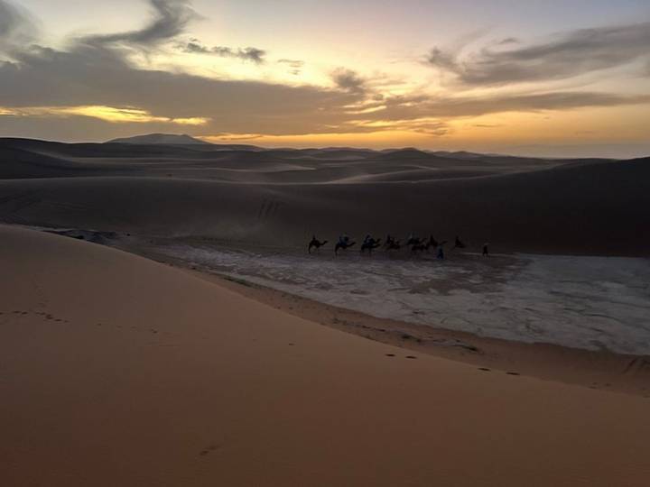 Camel caravan seen in the distance among darkening dunes as the sun sets behind the horizon.