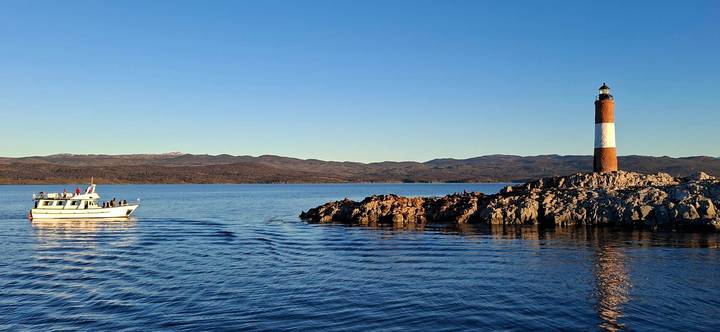 Red-and-white lighthouse on rocky islet with tourist boat on calm blue channel at golden hour.