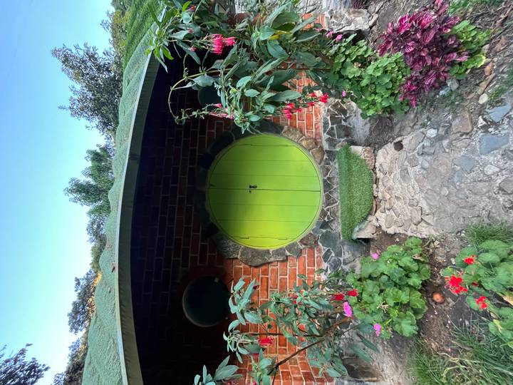 Small round green wooden door set into a brick and stone hut surrounded by garden flowers.