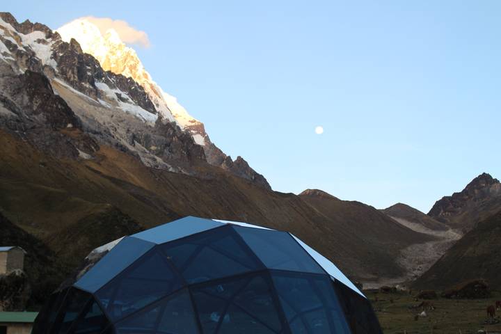 Geodesic glass dome campsite set against rugged mountains at dusk with the moon rising.