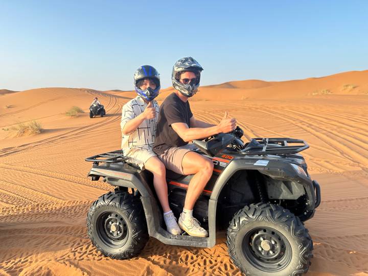 Two riders on an ATV give thumbs up while driving across golden desert dunes under a clear sky.