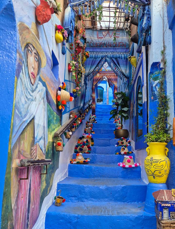 Vivid blue alleyway in Chefchaouen decorated with colorful woven hats and murals leading up painted stairs.
