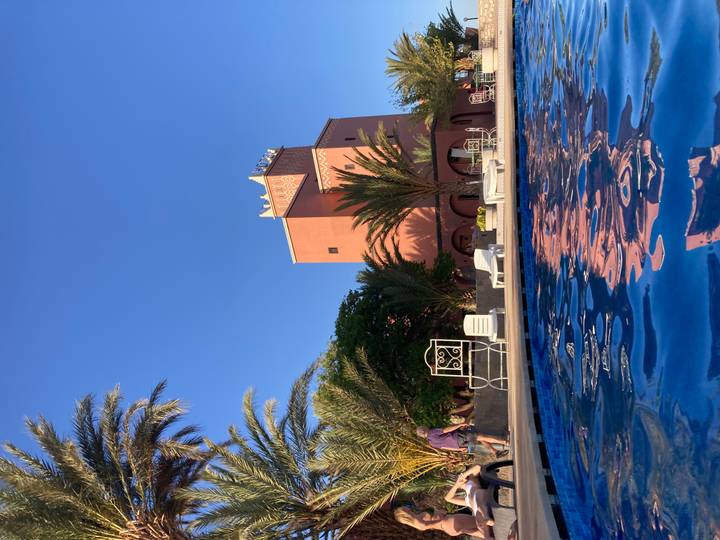 Swimming-pool perspective of a kasbah-style hotel tower with palm trees against a clear evening sky.