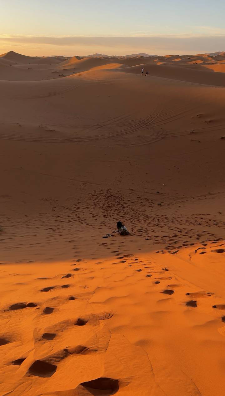 Lone traveler sits on the vast slope of a desert dune at dusk, footprints dotting the sand.