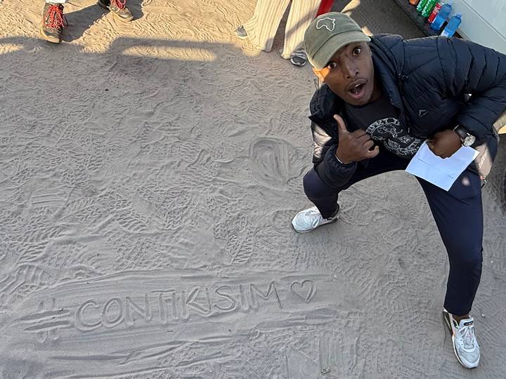 Traveler crouches on sandy ground posing beside the word "CONTIKISIM" drawn in the dirt.