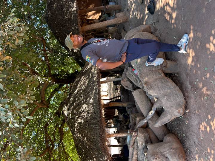 Traveler poses under shady trees next to traditional thatched huts in a cultural village setting.