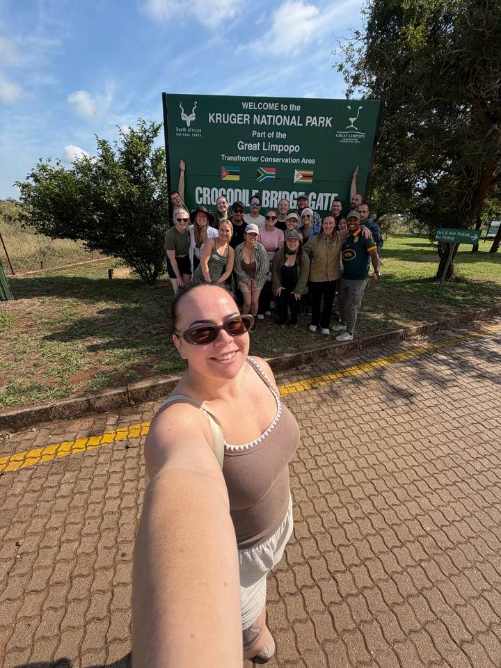 Selfie of a traveler with her tour group gathered near a green park sign on a sunny day.
