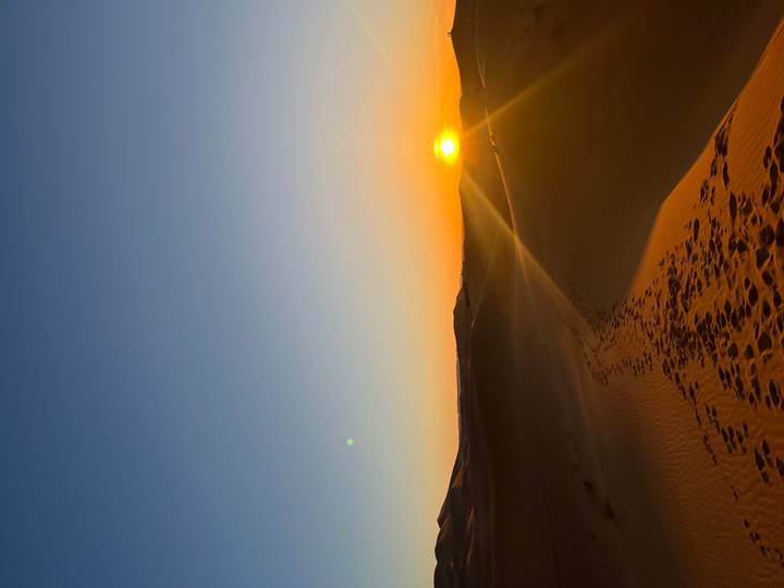 Rising sun glows over desert dunes with a trail of footprints leading toward the horizon.