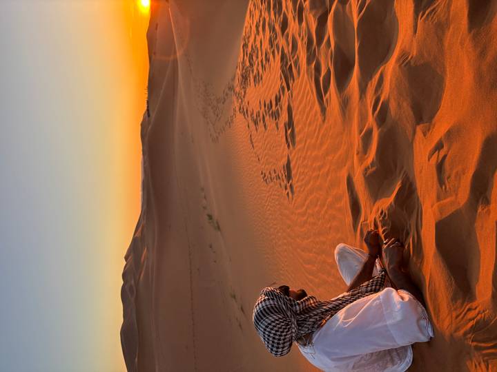 Man in traditional scarf sits on warm-lit dune watching the sun set over desert peaks.