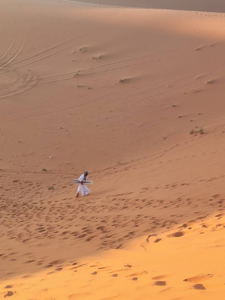 Local guide carries a sandboard up a steep dune, footprints trailing behind on the orange slope.