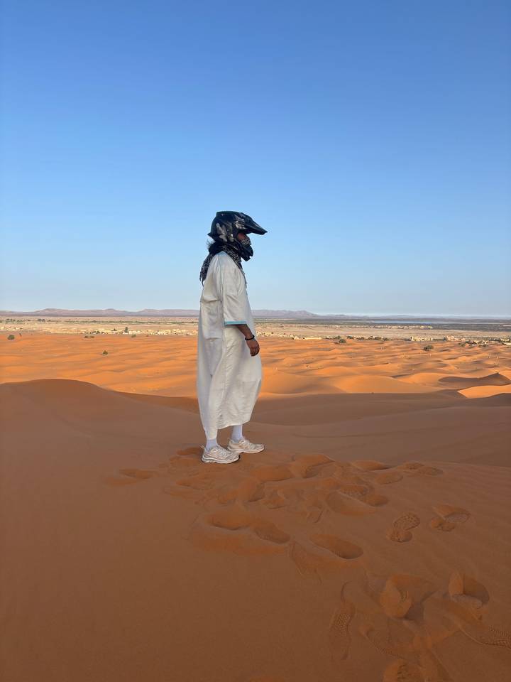 A lone traveler in a white robe and helmet stands atop orange sand dunes overlooking a vast desert landscape under a clear blue sky.