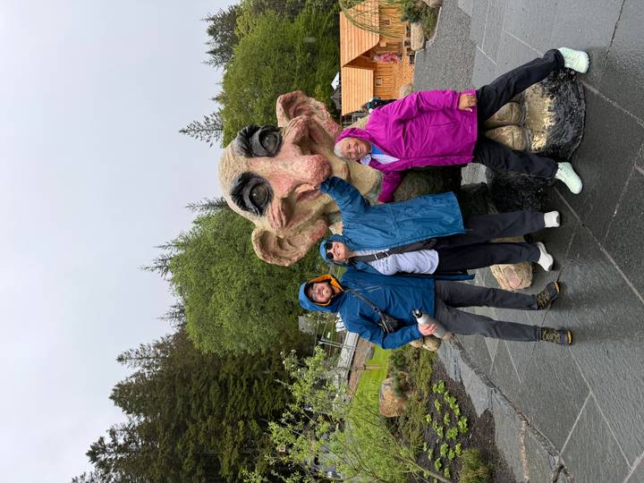Three travelers in rain jackets interact playfully with a large troll statue in a forested park.