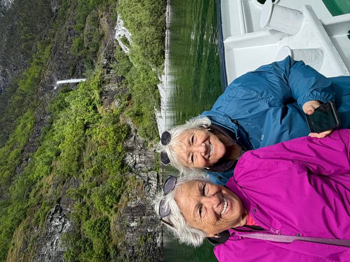 Two smiling women ride a boat through a lush fjord with a tall waterfall in the background.