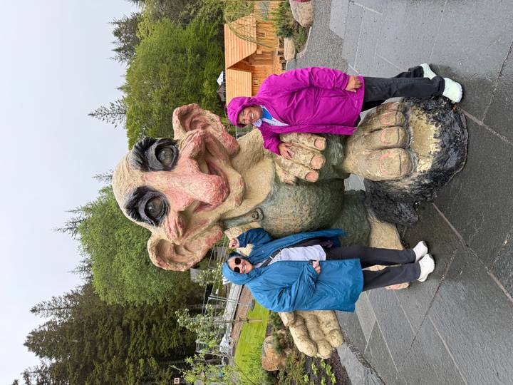 Two women pose with a large whimsical troll statue on a wet stone plaza.