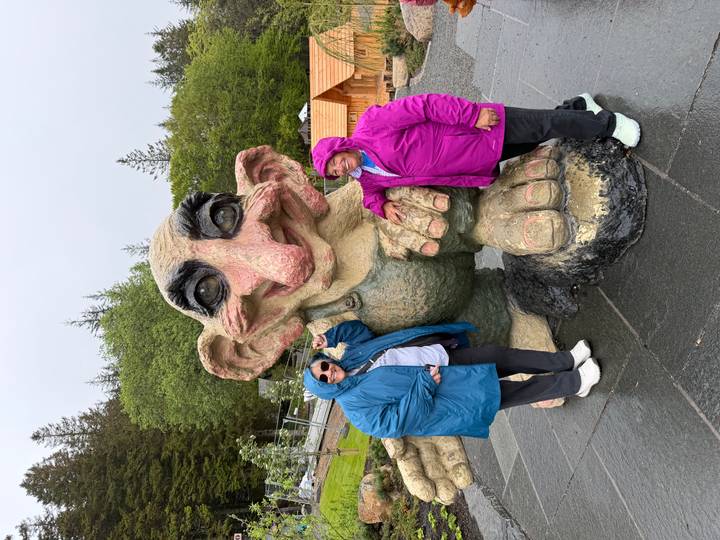 Two travelers in colorful rain jackets stand beside the nose of a giant troll sculpture.