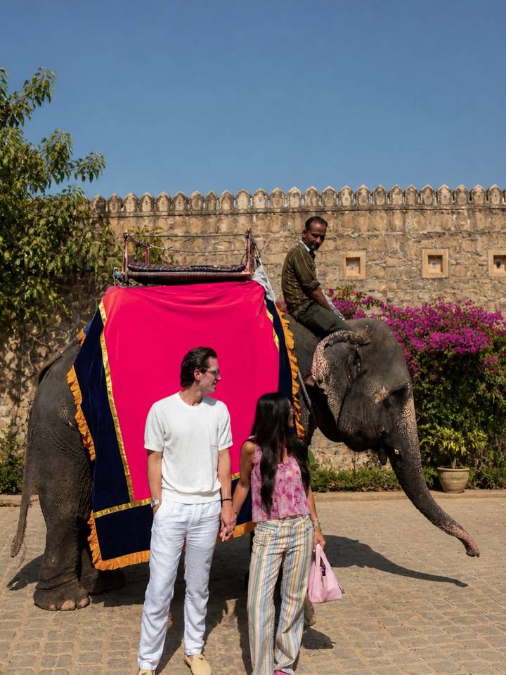 A couple stands before a brightly decorated elephant with a mahout seated on its back against an old stone wall.