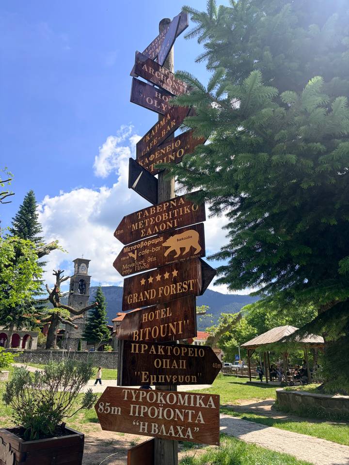 Wooden signposts in Greek point toward taverns, hotels, and forest hikes under a partly cloudy sky.