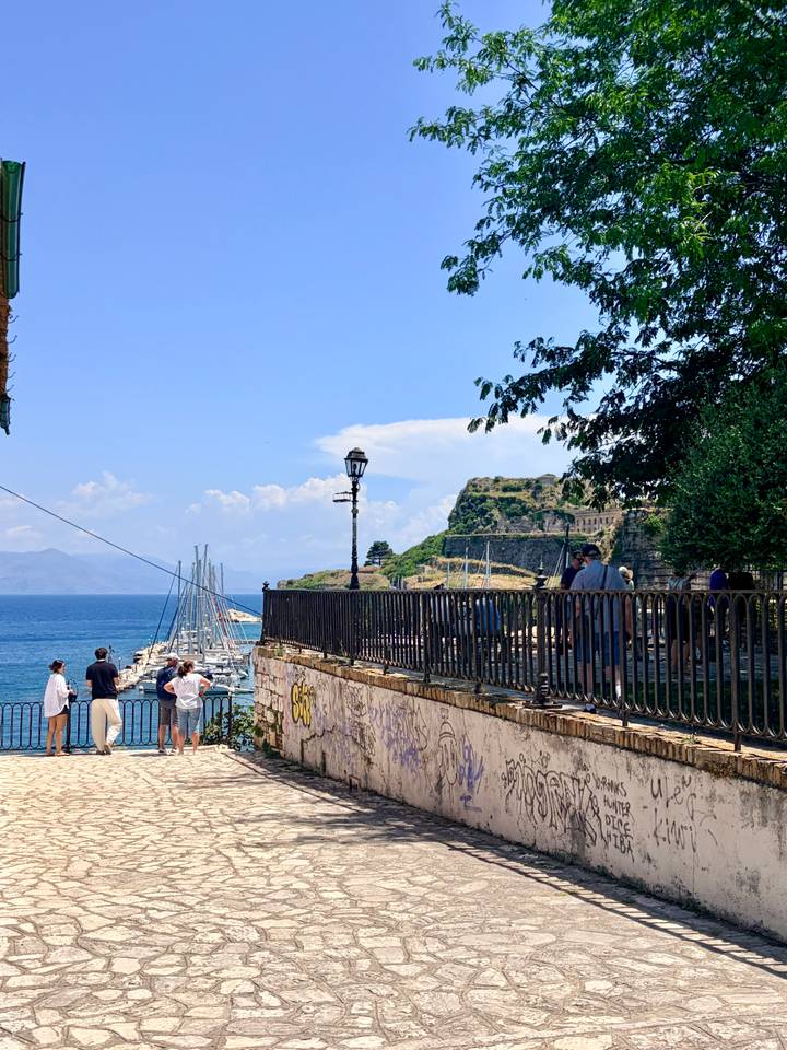 Visitors admire a marina and fortress walls overlooking the blue Ionian Sea on a sunny day.