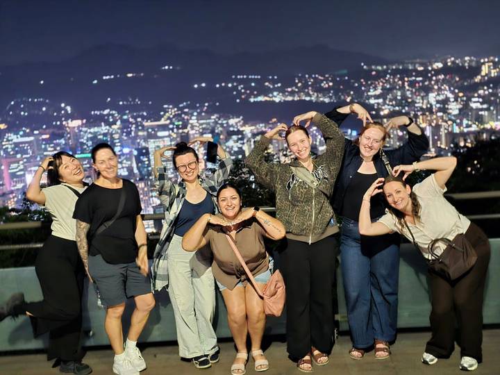 Group forms heart shapes with arms against a sparkling night skyline of Seoul.