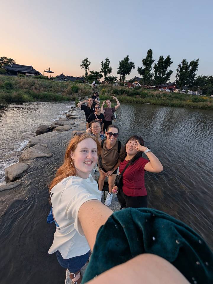 Friends laugh while crossing a river on stepping stones at dusk.