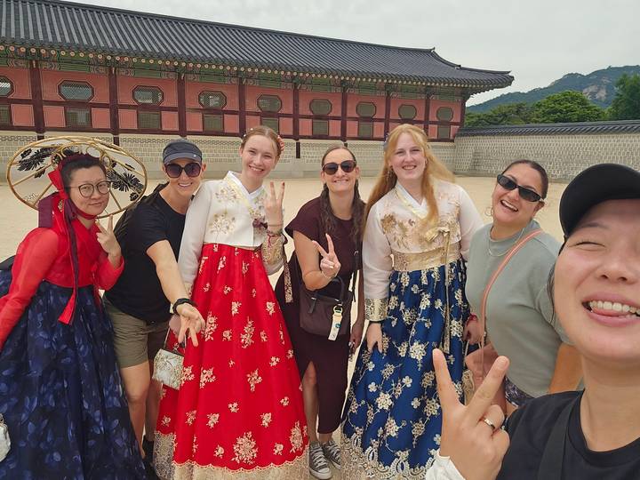 Travellers wearing colorful hanbok pose outside a palace gate in Seoul, flashing peace signs.