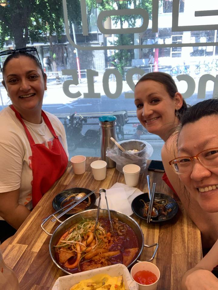 Three friends in aprons smile while enjoying noodle bowls at a casual eatery.