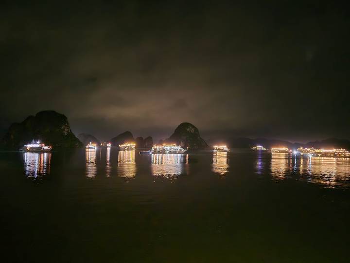 Night panorama of limestone karsts in Halong Bay with cruise boats lit up and reflections shimmering on calm water.