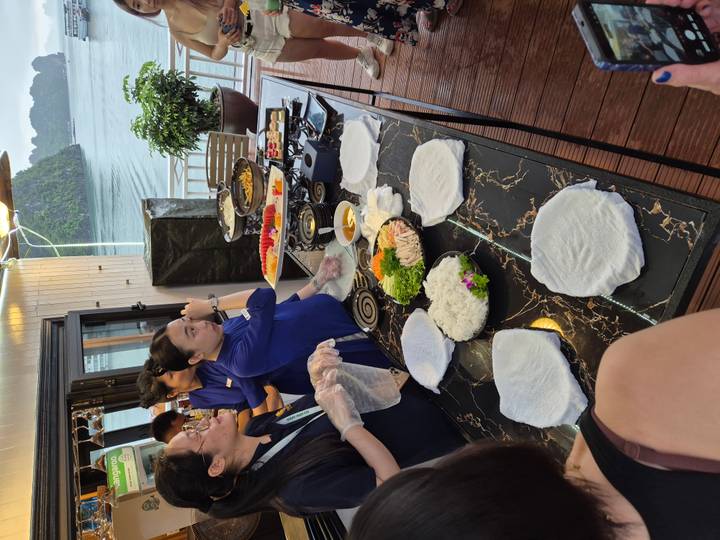 Cooking demonstration on a marble counter aboard a cruise boat, staff preparing fresh Vietnamese spring rolls with Halong Bay scenery in background.