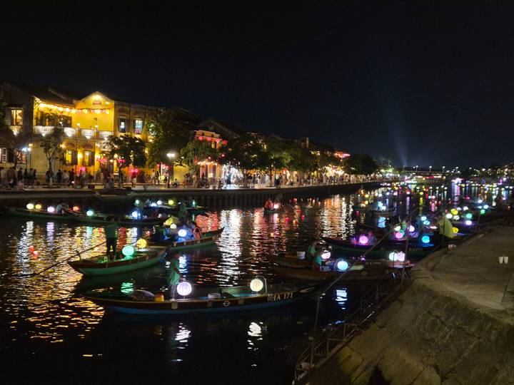 Hoi An riverside at night with colorful lantern-lit boats reflecting on the water and historic buildings glowing along the promenade.
