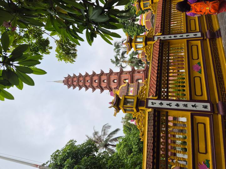 Tall red-brick pagoda rising behind an ornate yellow fence with tropical trees framing the scene.