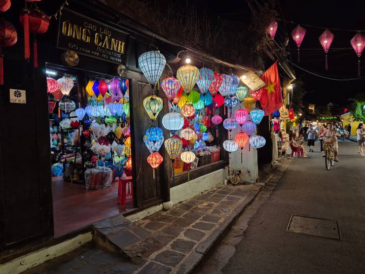 Night street in Hoi An decorated with dozens of vibrant silk lanterns outside a shop while pedestrians stroll by.