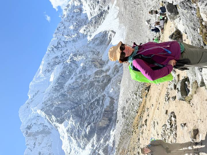 Female trekker smiles in front of a towering snow-capped Andean peak on a sunny day.