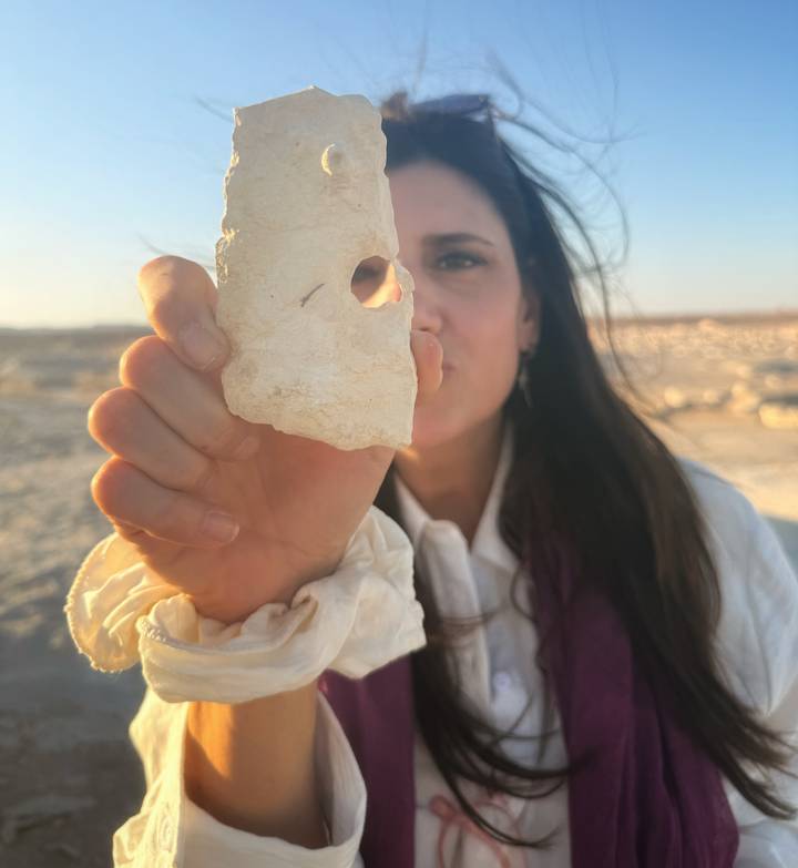Woman holding a pale desert rock fragment toward the camera with arid landscape behind