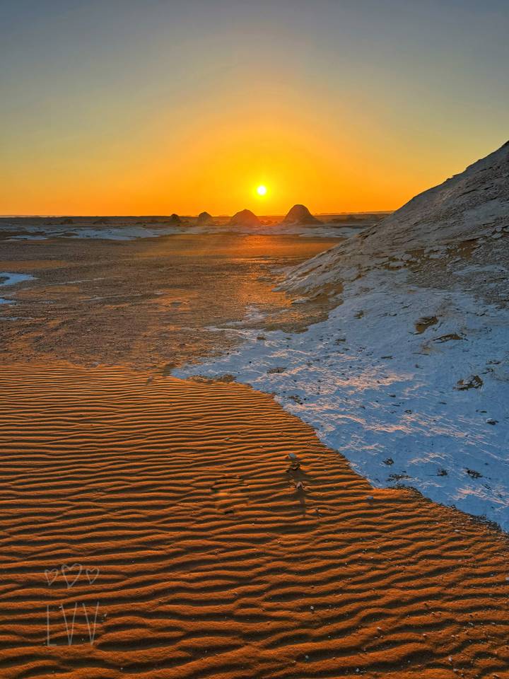 Rippled orange sand meeting pale chalk ridges with the sun setting behind distant formations