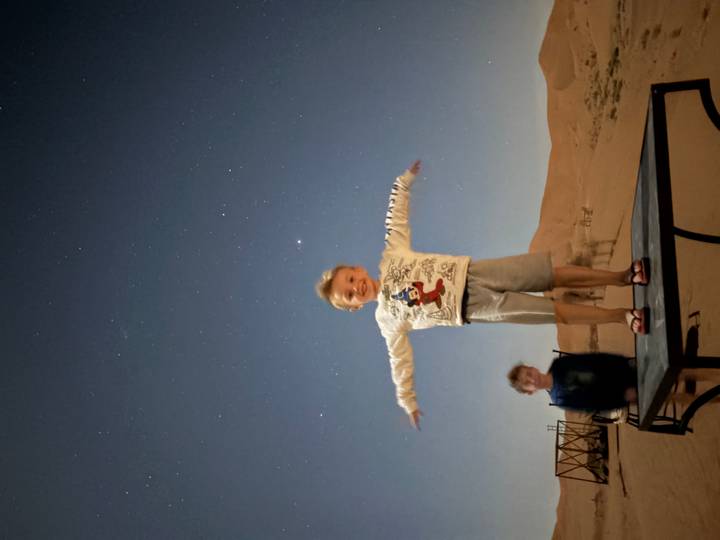 Young boy standing arms outstretched on a platform under starry desert sky, sibling blurred behind