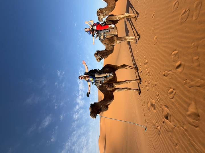 Travelers riding camels with arms raised against a blue desert sky