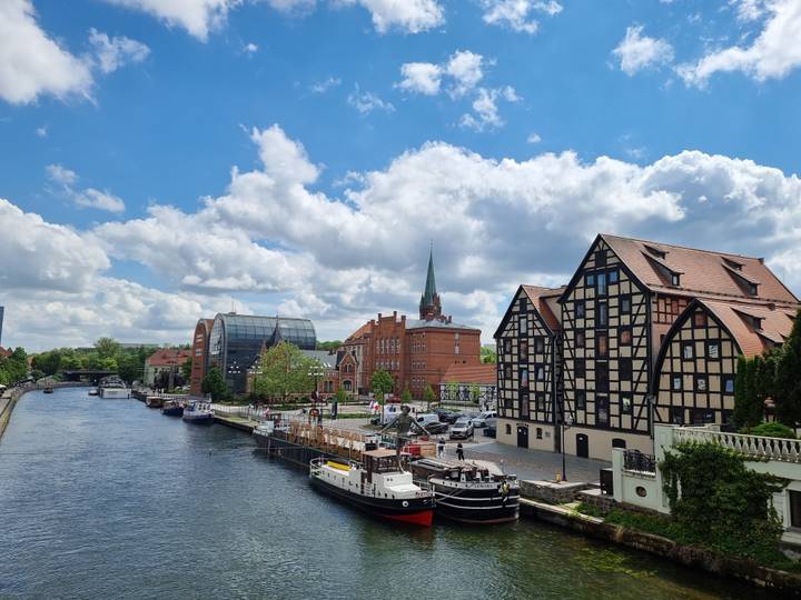 Charming riverside scene with half-timbered warehouses and moored boats beneath a bright sky.