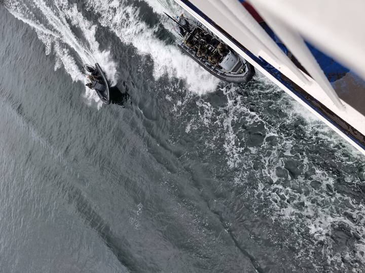 Motorized inflatables zipping alongside a large vessel on choppy grey water.