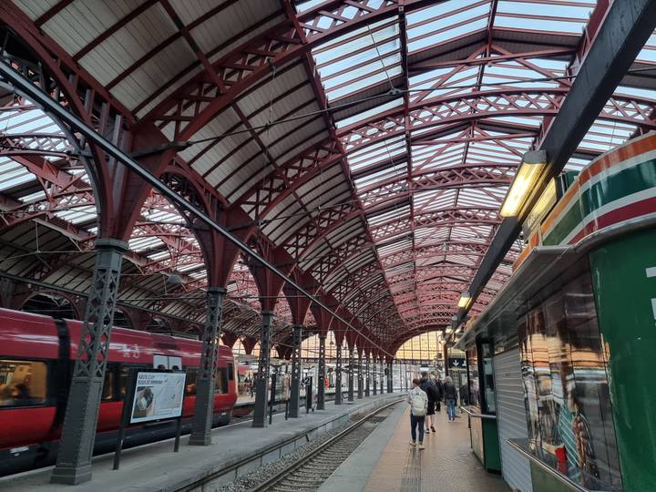 Interior of a historic iron-roofed railway station with red girders and waiting trains.