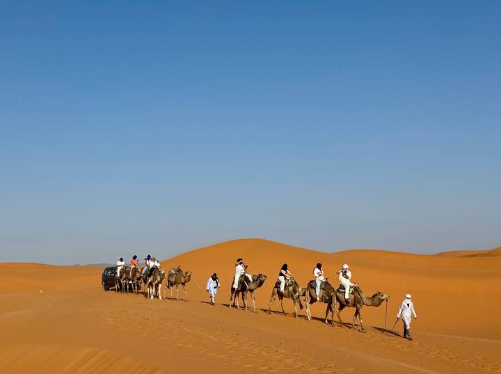 Caravan of riders and camels traversing bright orange Sahara sand under a cloudless sky.