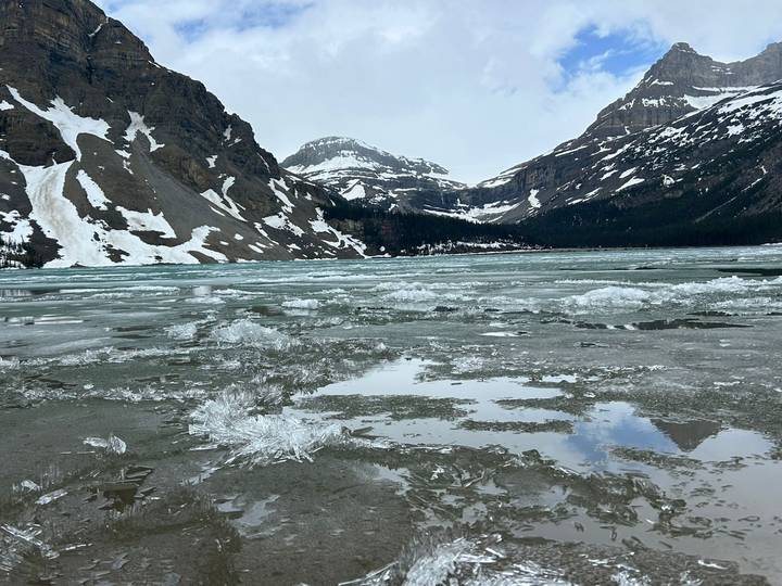 A partially frozen alpine lake with crystal ice shards, surrounded by snow-patched rocky mountains.