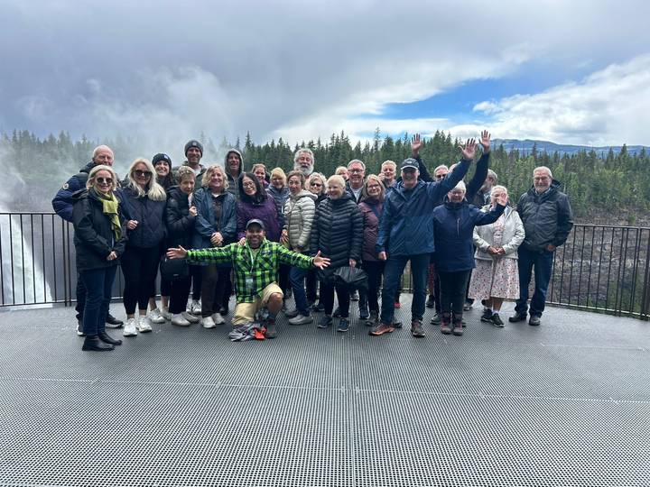 A large tour group in outdoor gear posing happily on a viewing platform beside a misty waterfall and forest.