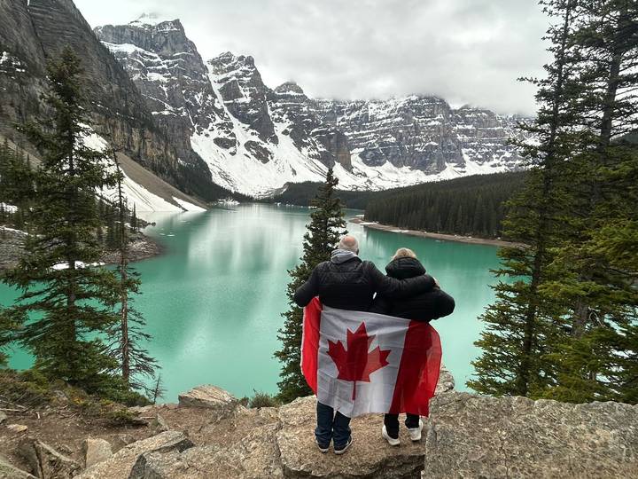 A couple wrapped in a Canadian flag gazing over the turquoise waters of Moraine Lake amid snow-capped peaks and evergreens.