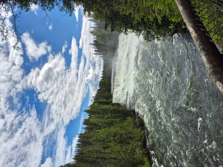 A powerful waterfall crashing through a forested canyon under a partly cloudy blue sky.