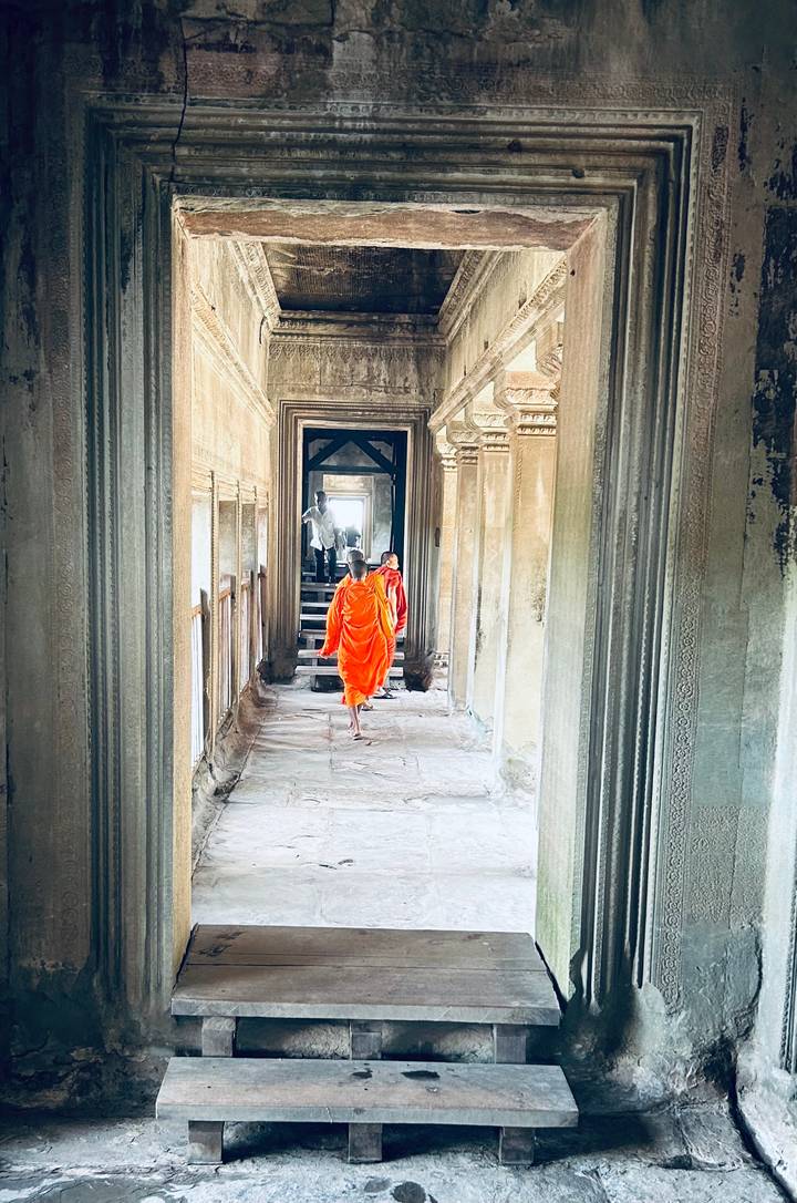 Buddhist monks in bright orange robes walk through a stone corridor of an ancient temple with detailed carvings.
