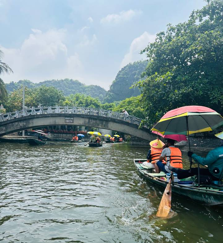 Tourists in small rowboats wearing life-jackets glide under a stone bridge amid limestone karst scenery.