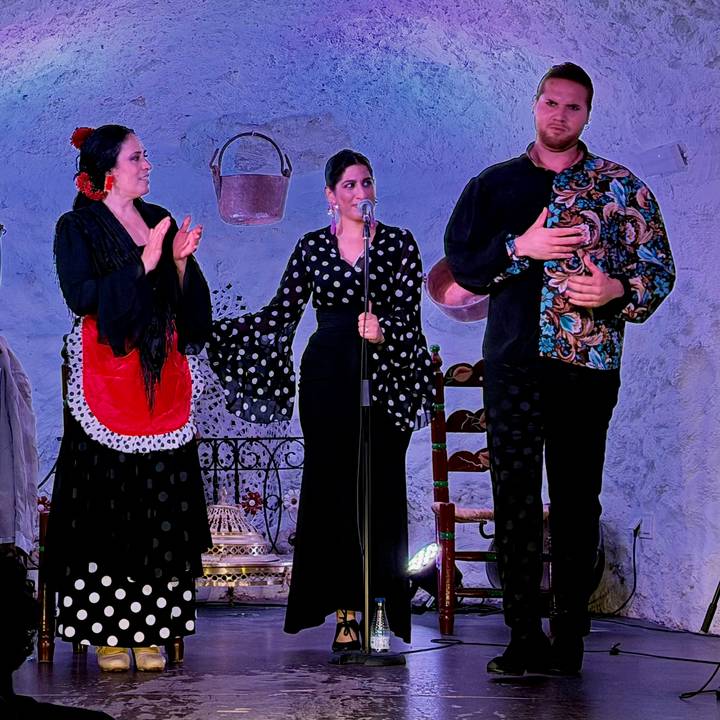Three flamenco performers on stage applauding the singer during a show under violet lighting.