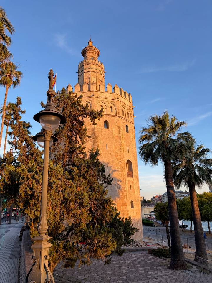 Golden-lit Torre del Oro flanked by palm trees and old street lamp.