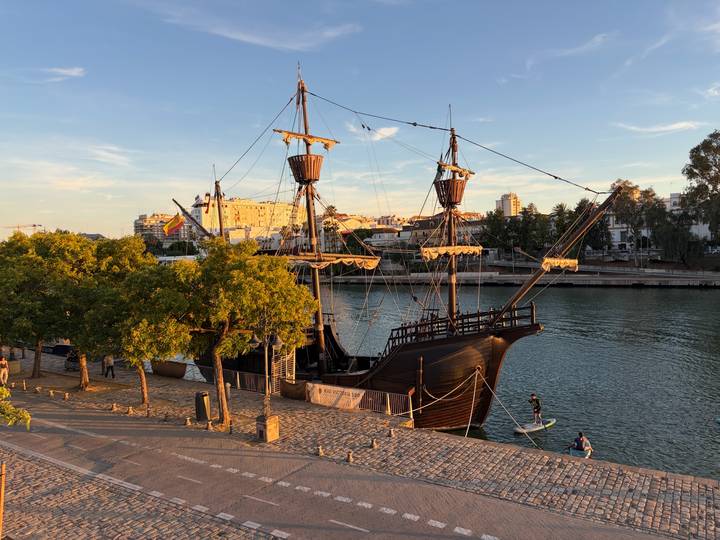 Replica wooden galleon moored on a calm river with paddle-boarder nearby at sunset.